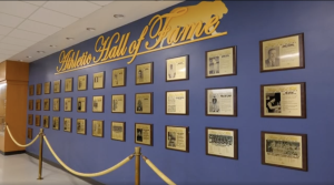A wall display featuring numerous gold-framed plaques honoring inductees in the COC Athletics Hall of Fame. The wall is painted blue with "Athletic Hall of Fame" written in large, elegant gold lettering at the top, alongside the outline of a mascot or figure.
